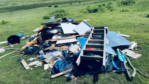 The pile of fly tipping waste at Cadover. The pile of rubbish includes a wooden ladder, wood and a plastic mannequin. The was has been left on a green grass area.