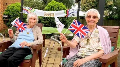 Two ladies smiling at the camera. They are both holding Union flags and Olympic flags. They are sitting on wooden deckchairs on a paved area in a garden. Behind them is a banner that reads "Go for Gold 2024" in gold lettering