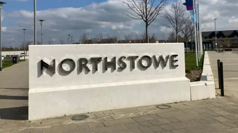 Emma Howgego/BBC A large white stone has the words 'Northstowe' written on it in dark shiny letters. To the right are some flag poles with flags and some new build buildings in the background. 