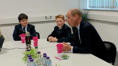 A photo of Prince William speaking to two students. They are speaking around a table with pink cups in the middle of the white table.