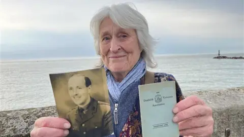 Phil Harrison/BBC A woman with white hair smiling at the camera and standing by a wall with the sea behind her. She is holding a black and white picture of a man and a small book which says "Dunkirk Veterans Association" on the front.