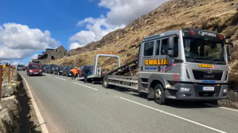 It's pen y pass road. On the right there is a lorry which with workers trying to lift a car which is illegally parked on to it. Behind the lorry there's a row of several other cars parked on the side of the road. There's also a red car driving past the parked cars.