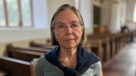 Martin Giles/BBC Jo Wareham is standing in the church, with rows of wooden pews behind her. She is looking directly at the camera. She is wearing a grey short-sleeved T-shirt with a navy cardigan wrapped round her neck.