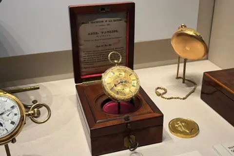 An elaborate gold pocket watch displayed with a brown display box in a cabinet