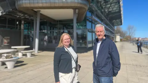 Carolyn Basings and Nigel Taylor from Save the National Glass Centre campaign smiling in front of the sun-lit venue. The multy-storey building is mostly made of glass and grey panels. It is by the river Wear with a waman strolling down the promenade in the distance. Carolyn has long, straight, blonde hair. She is wearing glasses, white trousers, navy-blue cardigan and a white blouse with blue graphics. Nigel is wearing jeans, navy blue jacket and a white t-shirt. He has short grey hair. 