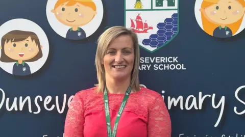 Leanne Smyth is wearing a pink top with lace sleeves and has a green lanyard around her neck. She is standing in front of the sign for Dunseverick Primary School which displays a school crest and cartoon-style portraits of pupils: two bots and two girls.