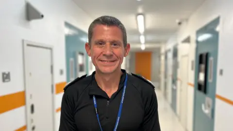 BBC/ Kate Bradbrook Chief constable Ivan Balhatchet stands in a bright corridor with doors on either side, wearing a black polo shirt and a blue lanyard around this neck smiling at the camera.