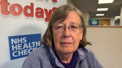 BBC A woman wearing glasses in a blue fleece in front of an NHS sign