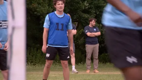 Football players train on a pitch with the club owner in the background, a man in brown trousers and navy blue top