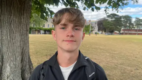 A teenager with short light brown hair wearing a white t-shirt and black anorak with a rucksack on his back with a school rugby pitch behind him.