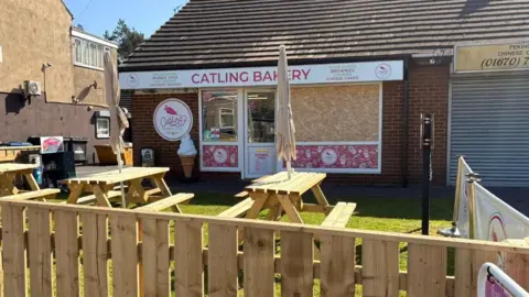 Catling Bakery Catling Bakery. The window has been boarded up with chipboard. Three picnic benches stand outside the shop.