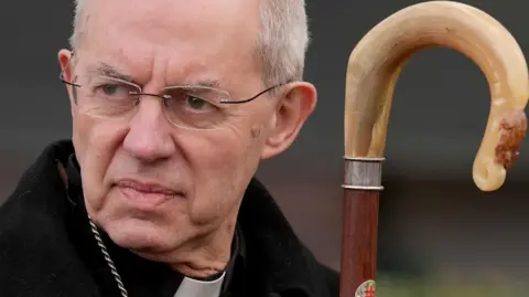 PA Media Justin Welby the archbishop of Canterbury looks to the left of frame. He's dressed in a black coat, and is wearing a priests dogcollar. 