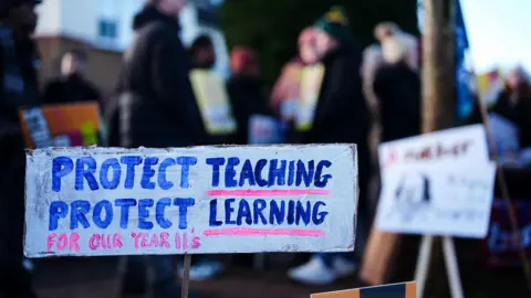 Signs belonging to members of the National Education Union on a picket line outside the Arthur Terry Learning Partnership (ATLP) school in Sutton Coldfield. The image shows a white rectangular sign made out of cardboard. The sign is painted with blue and pink lettering which reads: "Protect teaching, protect learning for our Year 11s." 
