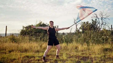 A person in a black vest and shorts stands in a field waving a trans flag.