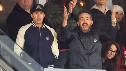 Rob McElhenney [left] and Ryan Reynolds [centre] in the stands watching Wrexham