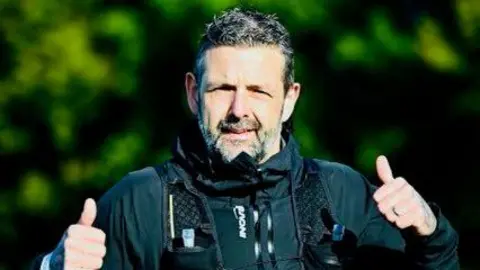David Graham A man with short dark and grey hair and stubble is doing a double thumbs up. He is wearing a black, outdoor running outfit, including a running vest and lightweight jacket. 