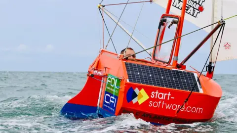 Andrew Bedwell Andrew Bedwell, in his early 50s, peeks out from inside a tiny orange yacht on the open ocean, which has a white sail and is covered with signs for various sponsors. He is wearing a black life vest and red waterproofs.