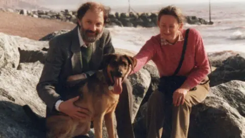 Dressed in a brown suit Leon Garner sits on the rocks by the sea at Sidmouth and smiles at the camera. He has his hand around his large dog Jenny. his wife Sandra is sitting on another rock close by and she is also patting the dog. She is wearing a pink jumper and brown trousers and the sea can be seen against the shore behind them. The dog is panting and has her tongue out.