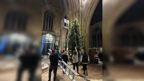 A number of volunteers, one holding a ladder and one in the top of a cherry picker decorate the tree which has been restored to its upright position