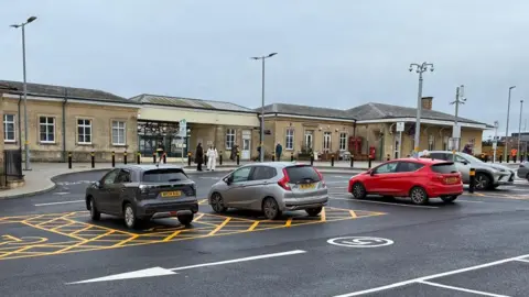 A car park with several cars parked. In the backgound is a station people with people walking past.