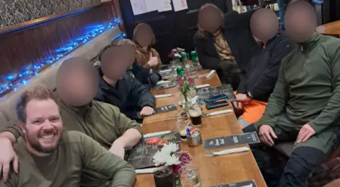 The Homeland Party A group of eight people sitting around a table in a pub. They are all looking at the camera. All faces have been blurred except for David Gardner, in the bottom left of the photo. He is bearded and smiling at the camera. 