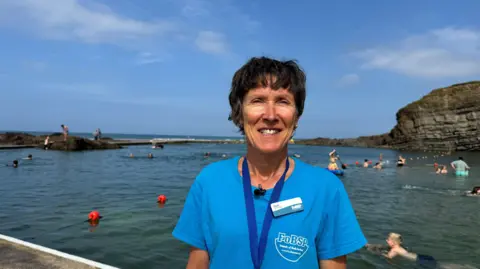 Deb Rosser smiles at the camera and stands in front of the sea pool which is busy with swimmers on a sunny day.