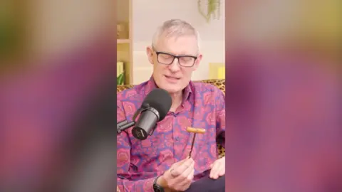 Jeremy Vine holding a gimlet tool. He's wearing a red and blue shirt and black framed glasses.