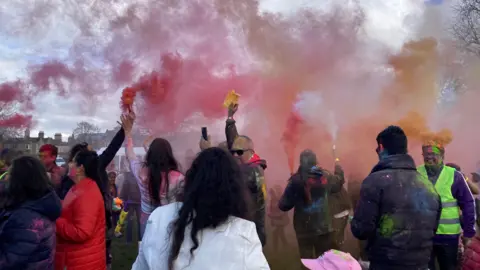 A crowd of people, their faces and clothes covered with multi-coloured bright paint, hold paint cannons in the air. Red and orange hues fill the air. Everyone is smiling and standing in small groups. 