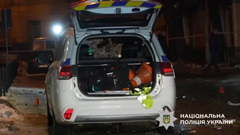 National Police of Ukraine/Telegram A white police car with its boot open pictured in Lviv at night. Its windshield is shattered and debris following the blasts is visible.