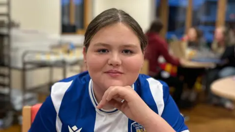 Rhianna Venables/BBC Scarlett is wearing the blue and white striped Colchester United kit. She is leaning on a hand and smiling. She has brown hair and is sitting on a chair.