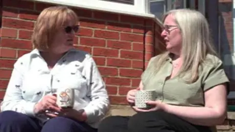 The two friends Amelia and Nadeen are sitting drinking tea in a garden in deep conversation. It is sunny and there is a brick wall behind them.