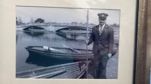 BBC/Unknown A black and white photo of Mac in a full suit and hat standing next to a lake, bridge and boats in East Park