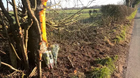 BBC The side of a rural road showing bunches of flowers propped up against a tree. The hedge to the right, between the road and a field, appears to have been damaged.