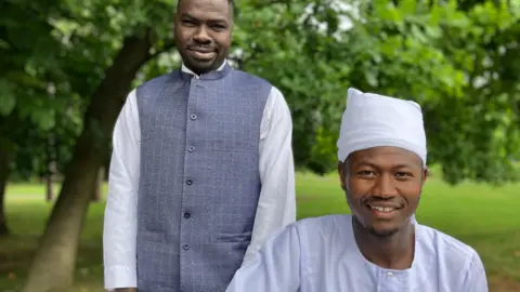 Jon Wright/BBC Two men in formal clothes, one with a white headscarf and tunic, smile at the camera with trees and grass behind.