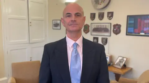 Mark Ansell/BBC A man wearing a blue suit, pink shirt and blue tie looking directly towards the camera