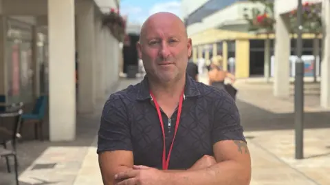 LDRS A bald man with a short beard stands with his arms crossed. He has a neutral expression and wears a collared navy t-shirt.