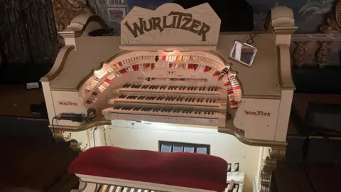 A view of the Wurlitzer organ in the Blackpool Tower Ballroom. It is white with the word Wurlitzer spelled out in big gold lettering on the music stand above three keyboards.