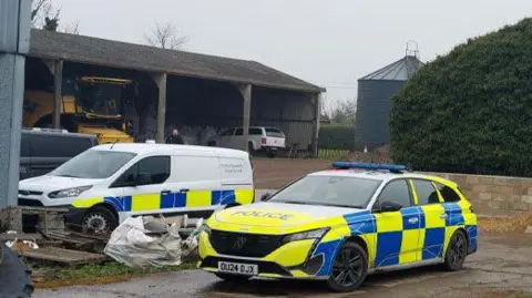 Cambridgeshire Police Three police vehicles - a van, an estate car and a four-wheel drive - parked at a farmyard. There is a barn to the left, a grain silo and a large tree or shrub behind the vehicles. 