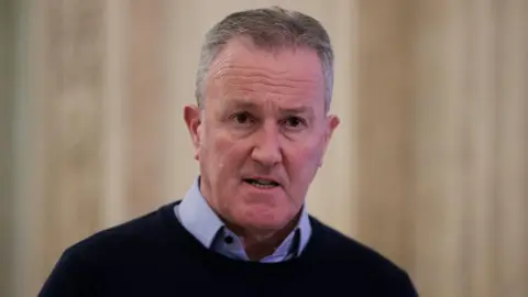 PA Media Conor Murphy - a man with short grey hair wears a dark jumper over the top of a blue collared shirt. He is standing in the main hall at Stormont, but the background is blurred.