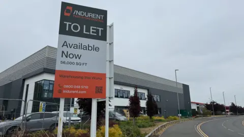 A sign including the words Indurent, to let and available now is prominent in the image in the foreground. A large grey facility including first-floor windows is in the background and part of a road is visible on the bottom right of the photo.