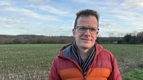 Martin Heath/BBC Simon Collyer with short dark hair and dark-framed glasses wearing a red and orange jacket and green and brown scarf. He is standing in a field which appears to have a crop planted in it. There are hedges and trees in the distance.