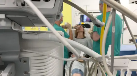 The picture shows the back of hospital equipment, the leads and wires are in focus and then in the background you can see a patient sitting while to surgeons work with a camera. they are slightly blurred.