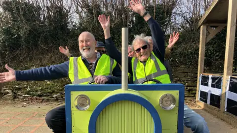 A group of men wearing high viz jackets and waving their hands in the air as if they are flying left on the wooden tractor. 