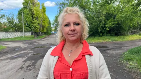 A woman with blonde hair stands at on a run-down intersection of a road. She wears a pale woollen cardigan, bright red shirt and a cross necklace.