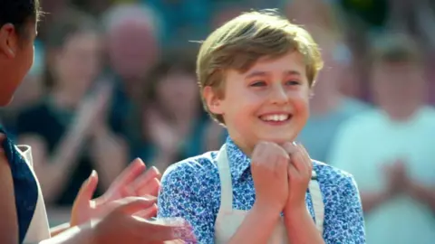 Channel 4/Love Productions 11-year-old Austin smiles and brings his hands to his mouth as he reacts to being crowned the winner. He is wearing a beige apron over a blue flowery shirt, and his straight hair is brushed to the side. 