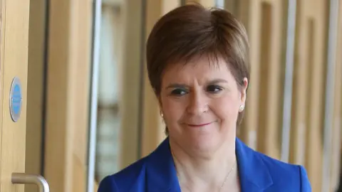 PA Media A close-up indoor photograph shows a Nicola Sturgeon who has short, neatly styled hair wearing a bright blue blazer over a light-coloured top and a round pendant necklace. She is standing near a wooden door with a visible metal handle and a small circular sign on the wall. The background consists of softly blurred wooden panels and glass, suggesting a modern interior space with natural light.