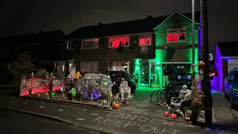 PA Media Their home in the dark. The path in front of the house says 'danger keep out' and their front garden is full of lit pumpkins, fake skeletons, cobwebs. The front door is lit green and the windows upstairs are lit red. 