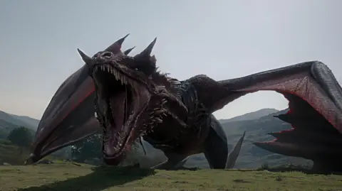 The head and neck of a large, red, computer-generated dragon rises over the top of the mountain; it is roaring, revealing sharp teeth with its wings outspread