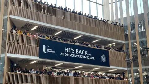 Crowds of people stand looking over wooden balconies in a large atrium in the Ulster University building. A huge navy UU-branded banner is draped across one of the balconies. It reads: "He's back, as Dr Schwarzenegger. 