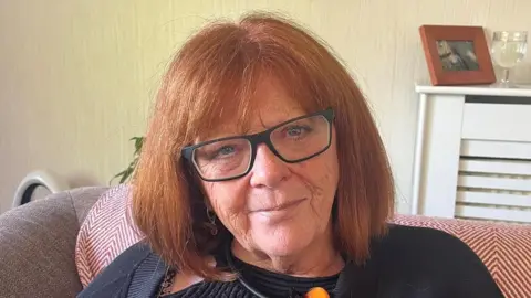 Tricia Donoghue has shoulder-length ginger hair with a fringe. She wears dark glasses and a black top and appears to be sitting on a couch in a living room.
